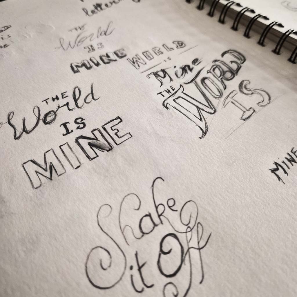 Hand lettering sketches in pencil, The World is Mine in several styles and Shake it Off in a flourished script