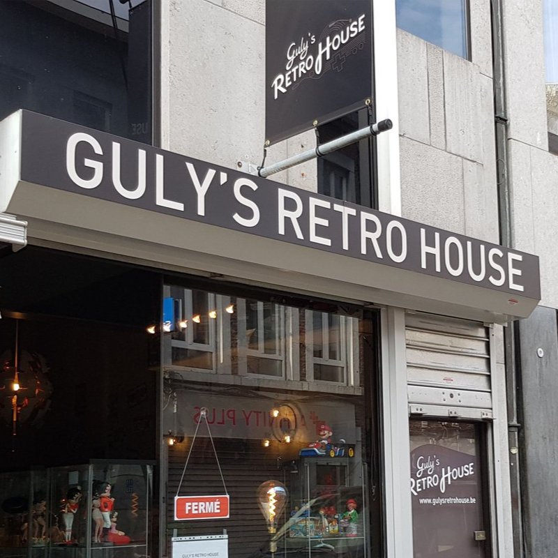 The shop front in Liège: the Retro House logo on the sign hanging over the street, the shop's name across the awning, figurines and a lamp in the window
